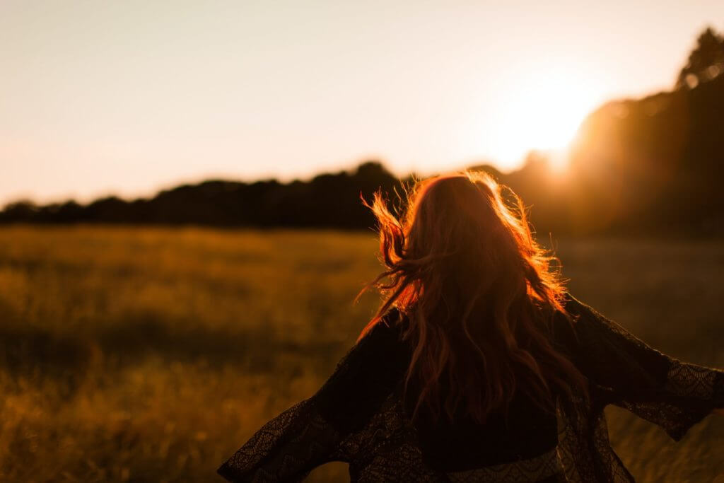 Une femme aux cheveux rouges debout dans un champ au coucher du soleil, réfléchissant à la façon de commenter réussir sa vie.