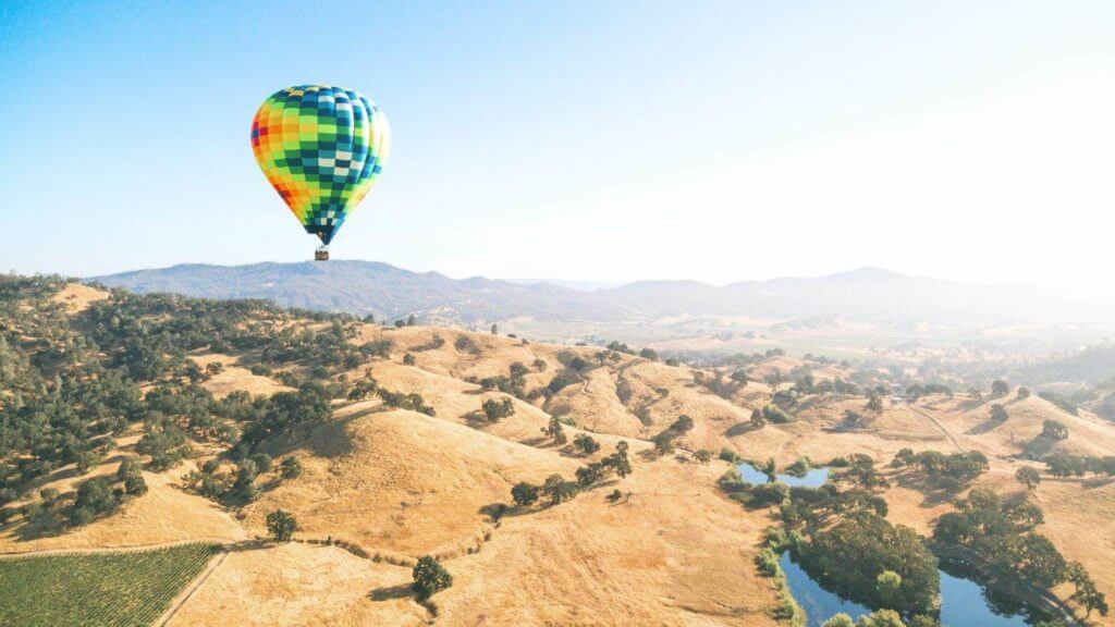 A colorful hot-air balloon flies over a field while passengers are mesmerized by the hypnotic views.