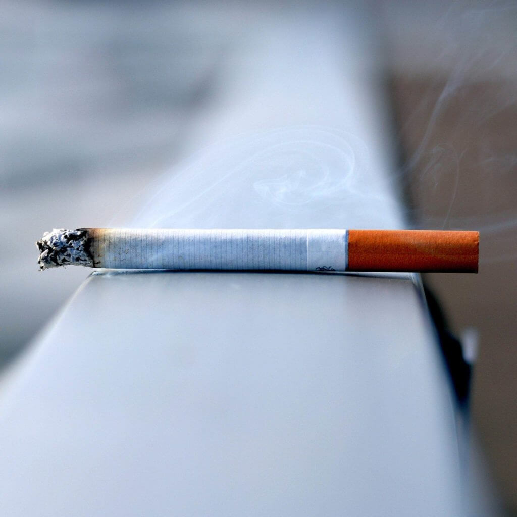 Une cigarette bien en vue sur une rampe métallique, émettant des volutes de fumée.