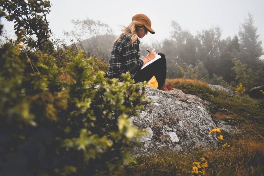 Une femme est assise sur un rocher et tient un journal dans un cahier.